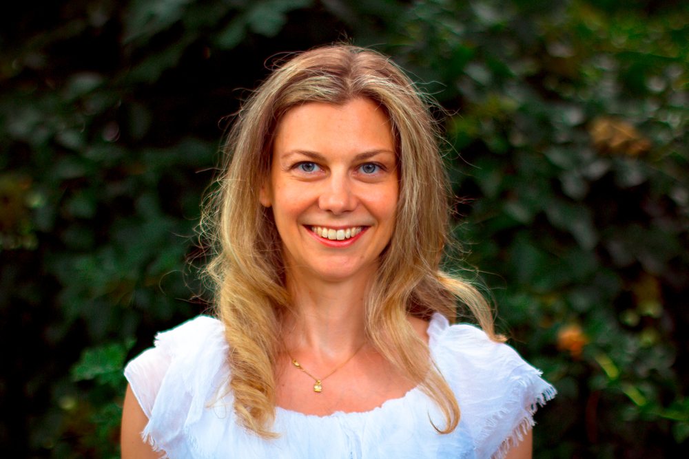 Julia Arbuckle in a white dress, smiling, with summer foliage behind her.